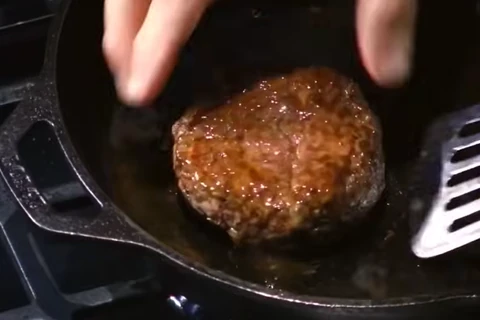 Searing seasoned beef patty in a hot cast iron skillet with a golden crust